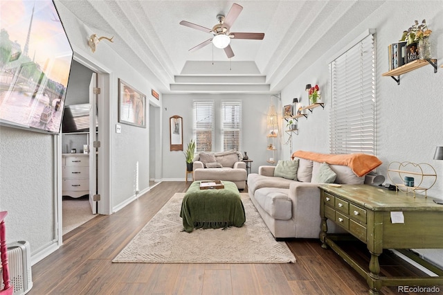 living room with a ceiling fan, baseboards, a tray ceiling, and dark wood-style flooring