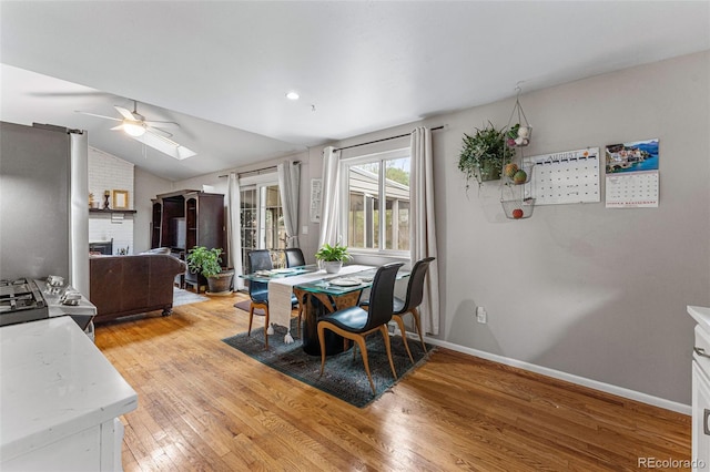 dining area with ceiling fan, vaulted ceiling, a brick fireplace, and light hardwood / wood-style floors