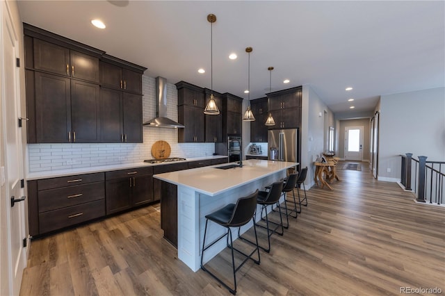 kitchen featuring appliances with stainless steel finishes, wall chimney exhaust hood, a kitchen bar, wood-type flooring, and a center island with sink