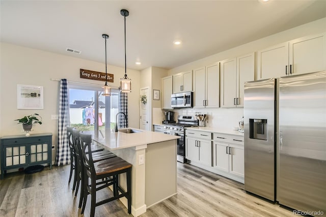 kitchen with pendant lighting, stainless steel appliances, light wood-type flooring, a kitchen island with sink, and sink