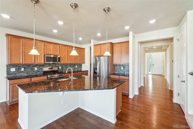 kitchen with brown cabinetry, a kitchen island with sink, a sink, dark wood-type flooring, and appliances with stainless steel finishes