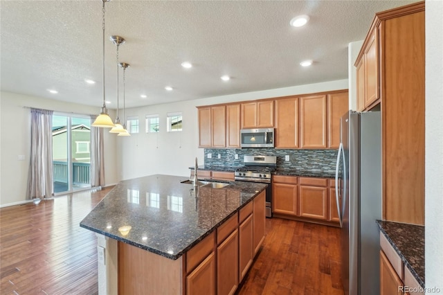 kitchen featuring dark wood-style floors, a center island with sink, a sink, decorative backsplash, and appliances with stainless steel finishes