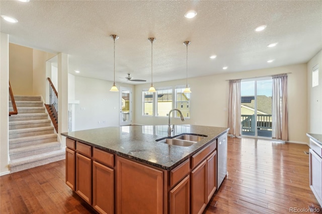 kitchen with dark wood-type flooring, an island with sink, a sink, dark stone countertops, and stainless steel dishwasher