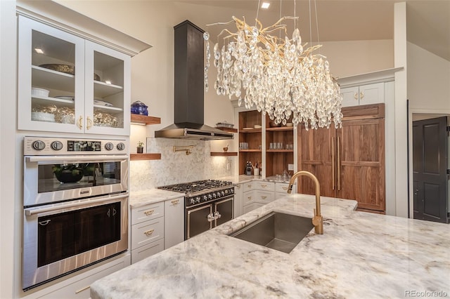 kitchen with a sink, high quality appliances, wall chimney range hood, light stone countertops, and glass insert cabinets