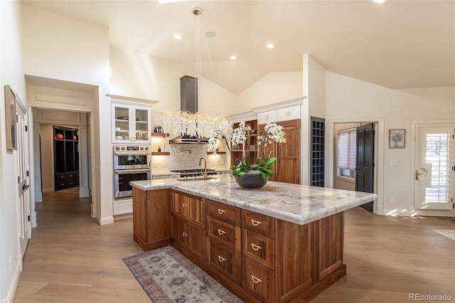 kitchen featuring glass insert cabinets, stainless steel double oven, decorative light fixtures, and an island with sink