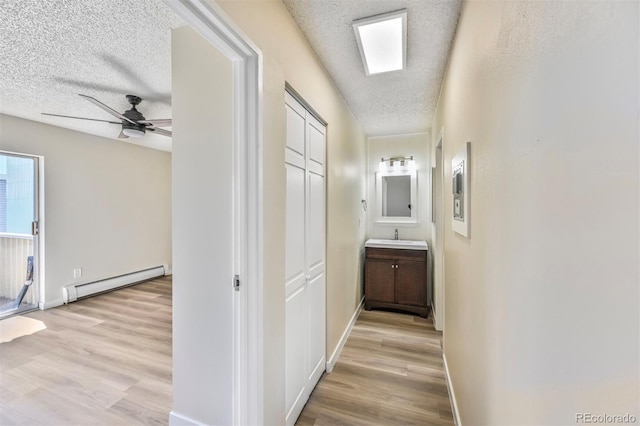 corridor with a baseboard heating unit, a textured ceiling, a sink, light wood-style floors, and baseboards