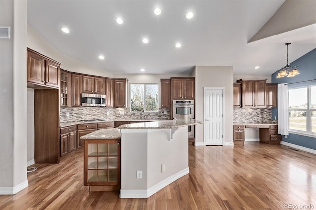 kitchen with light stone counters, hanging light fixtures, appliances with stainless steel finishes, a center island, and glass insert cabinets