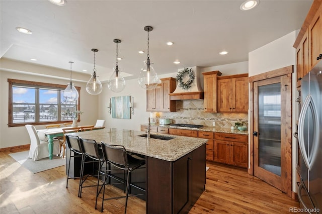 kitchen featuring pendant lighting, sink, premium range hood, appliances with stainless steel finishes, and a center island with sink