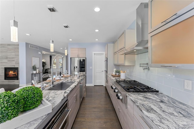 kitchen with appliances with stainless steel finishes, light stone counters, wall chimney exhaust hood, sink, and decorative light fixtures