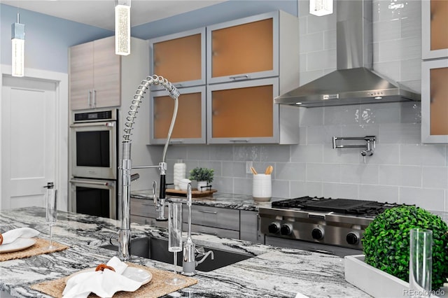 kitchen with light stone counters, wall chimney range hood, stainless steel appliances, and tasteful backsplash