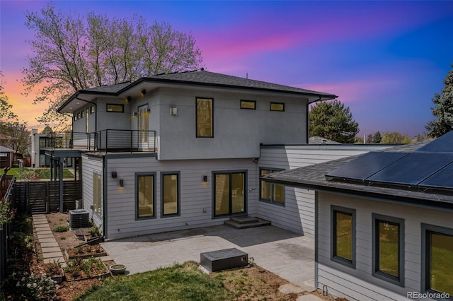 back house at dusk with a balcony, a patio, central AC unit, and solar panels