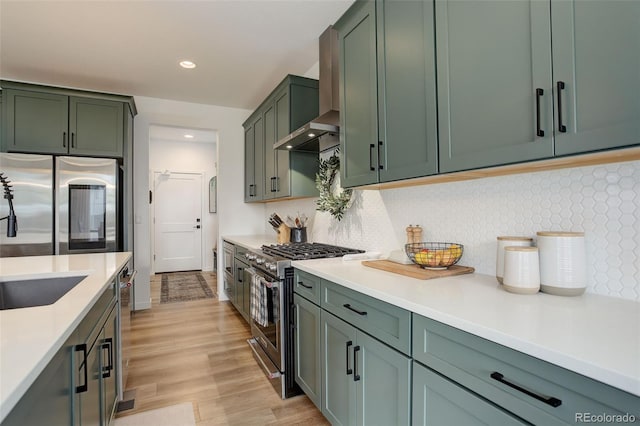 kitchen featuring light wood-type flooring, backsplash, green cabinetry, appliances with stainless steel finishes, and wall chimney range hood