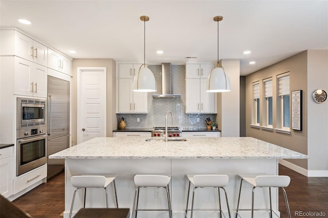 kitchen featuring built in appliances, wall chimney exhaust hood, an island with sink, light stone counters, and white cabinets