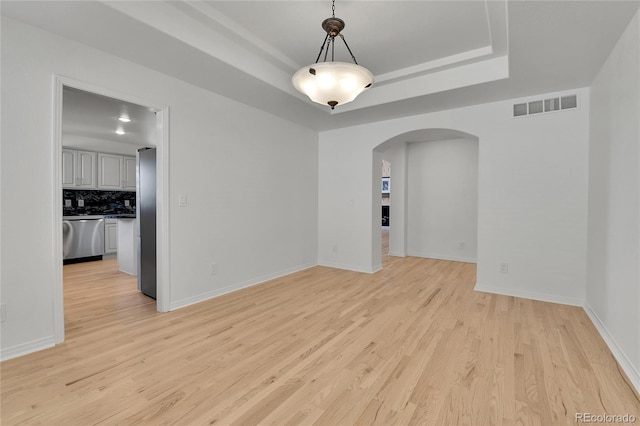 empty room with arched walkways, a tray ceiling, visible vents, and light wood-style floors