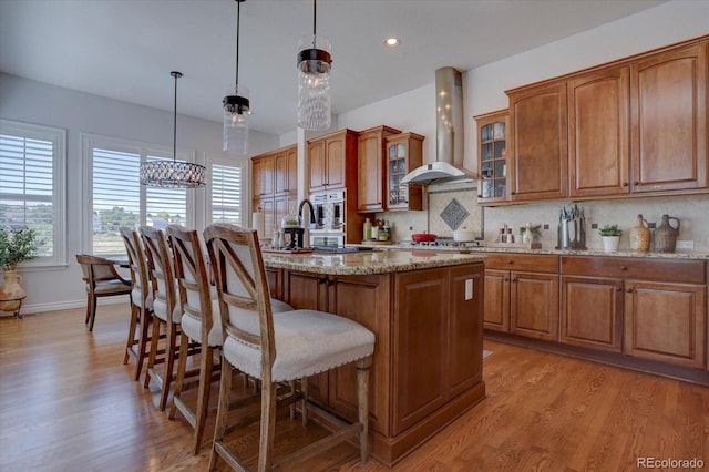 kitchen featuring backsplash, a kitchen island with sink, wall chimney range hood, hanging light fixtures, and light stone countertops