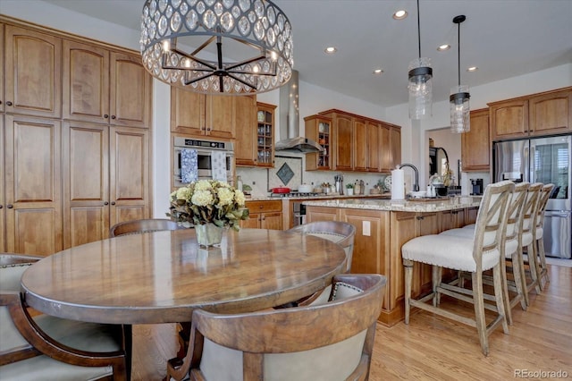 kitchen with stainless steel appliances, wall chimney range hood, decorative light fixtures, a kitchen island with sink, and light wood-type flooring