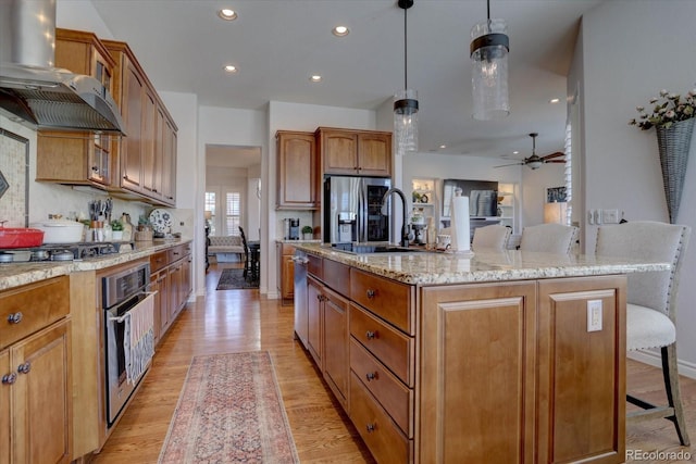 kitchen featuring hanging light fixtures, wall chimney range hood, an island with sink, appliances with stainless steel finishes, and light wood-type flooring