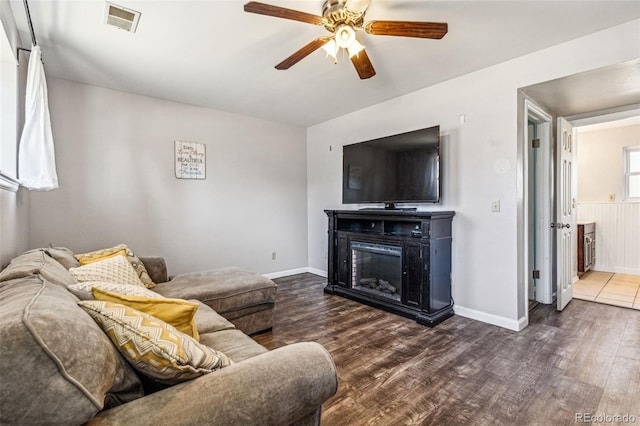 living area with a glass covered fireplace, dark wood-type flooring, visible vents, and ceiling fan