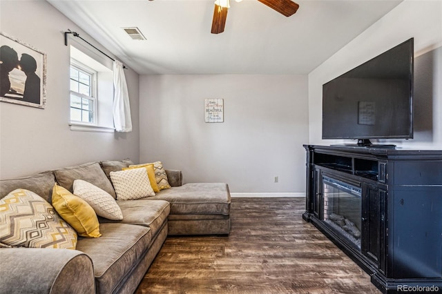 living area featuring visible vents, a ceiling fan, a glass covered fireplace, dark wood-style floors, and baseboards