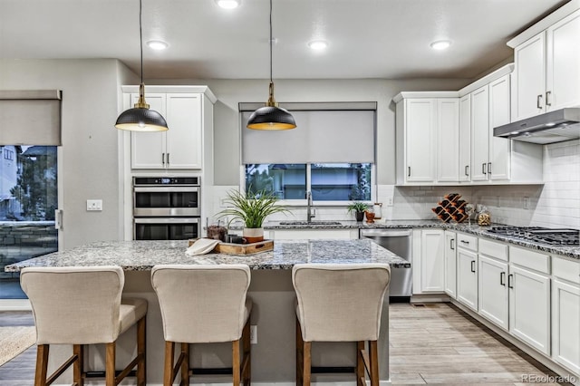 kitchen featuring light stone countertops, hanging light fixtures, stainless steel appliances, a kitchen island, and light wood-type flooring