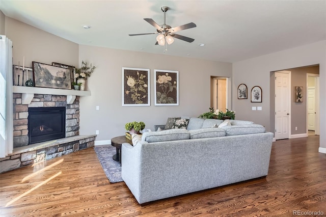 living area with ceiling fan, a stone fireplace, wood finished floors, and baseboards