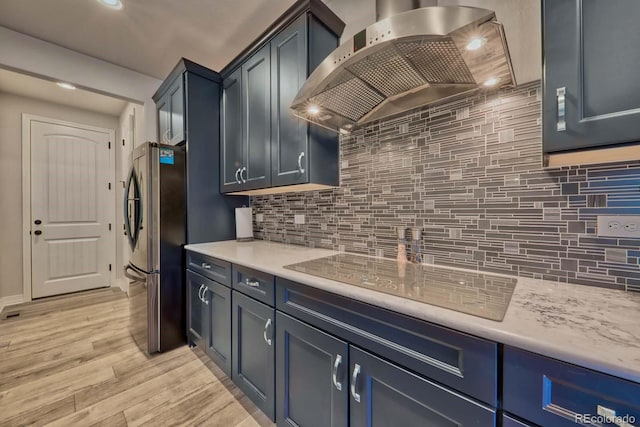 kitchen featuring backsplash, wall chimney range hood, stainless steel refrigerator, black electric stovetop, and light wood-type flooring