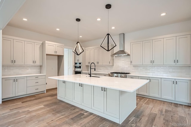 kitchen featuring decorative light fixtures, an island with sink, wall chimney range hood, appliances with stainless steel finishes, and sink