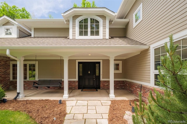 view of exterior entry featuring covered porch, roof with shingles, and brick siding