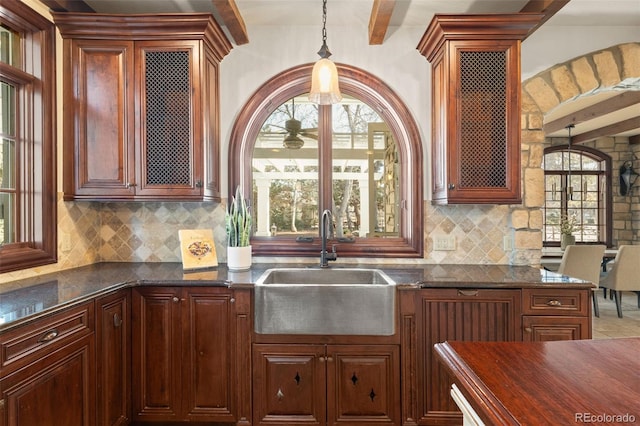 kitchen with reddish brown cabinets, decorative backsplash, decorative light fixtures, a sink, and beam ceiling