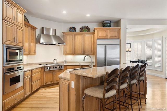 kitchen with a center island with sink, wall chimney exhaust hood, light wood-style flooring, built in appliances, and a sink