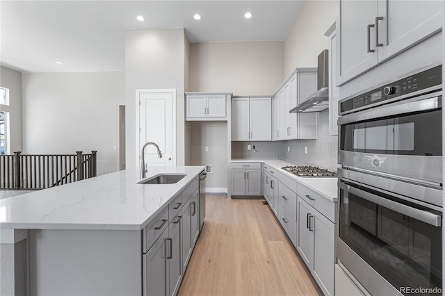 kitchen with light stone countertops, appliances with stainless steel finishes, an island with sink, sink, and wall chimney range hood