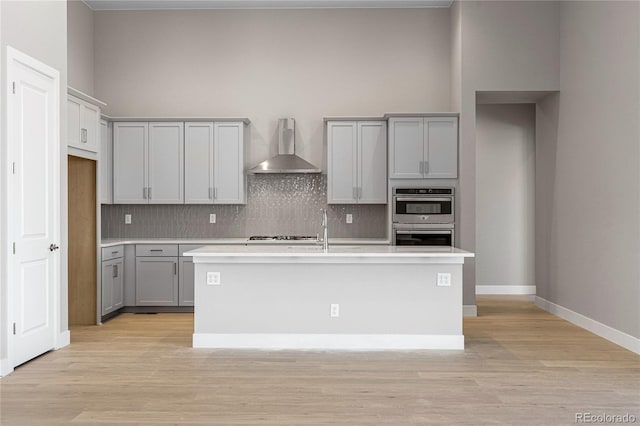 kitchen with stainless steel double oven, wall chimney exhaust hood, a center island with sink, and gray cabinetry