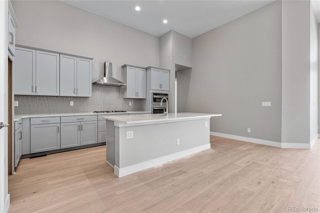 kitchen with wall chimney exhaust hood, a center island with sink, light wood-type flooring, gray cabinets, and backsplash