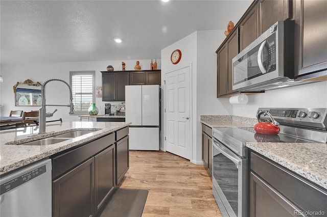 kitchen with dark brown cabinetry, appliances with stainless steel finishes, light stone countertops, a sink, and light wood finished floors