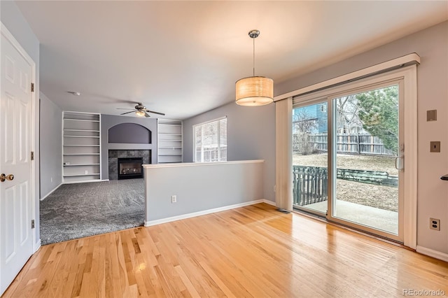 unfurnished living room with built in shelves, light wood-style flooring, a tiled fireplace, a ceiling fan, and baseboards