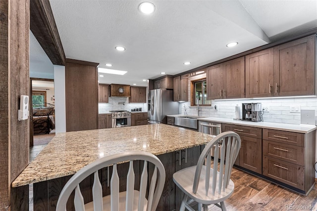 kitchen featuring dark wood-type flooring, appliances with stainless steel finishes, a kitchen breakfast bar, light stone countertops, and backsplash