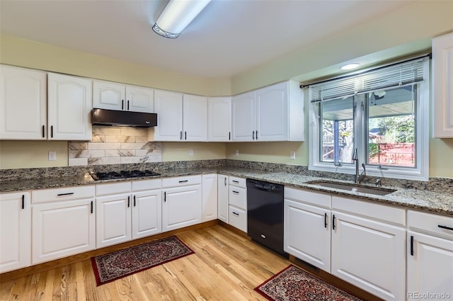 kitchen featuring black dishwasher, a sink, under cabinet range hood, dark stone counters, and light wood-style floors