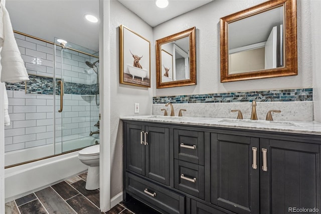 bathroom featuring wood tiled floor, double vanity, bath / shower combo with glass door, a sink, and toilet