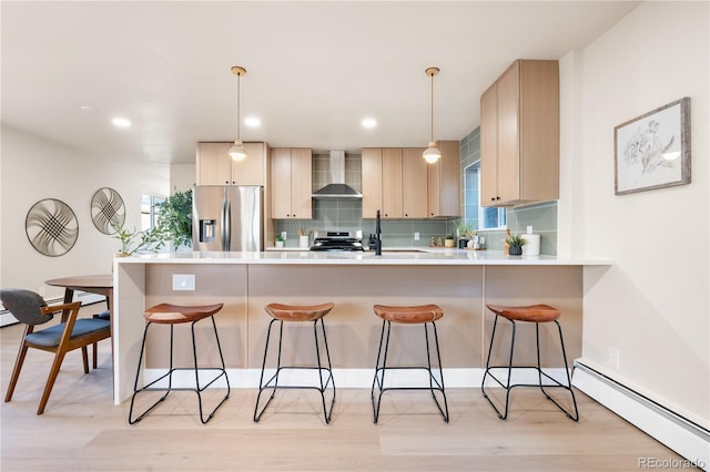 kitchen with appliances with stainless steel finishes, a baseboard heating unit, wall chimney range hood, light brown cabinets, and pendant lighting