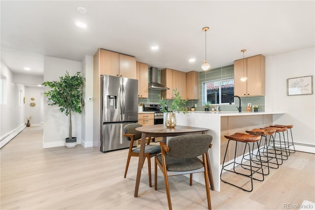 kitchen featuring kitchen peninsula, light brown cabinetry, wall chimney exhaust hood, stainless steel appliances, and pendant lighting