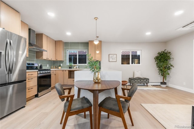 kitchen with light brown cabinetry, wall chimney exhaust hood, stainless steel appliances, light hardwood / wood-style flooring, and hanging light fixtures