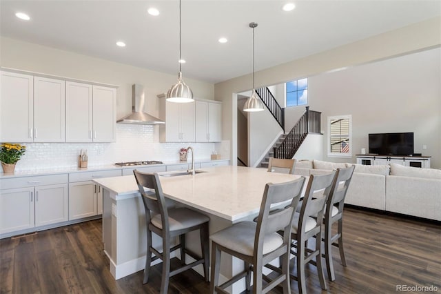 kitchen with a center island with sink, sink, hanging light fixtures, wall chimney exhaust hood, and white cabinetry