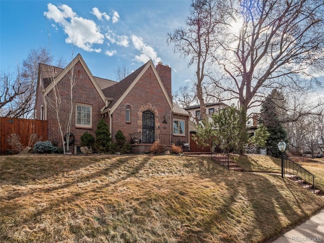 tudor-style house with brick siding, fence, a chimney, and a front lawn