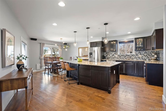 kitchen with a center island, stainless steel refrigerator with ice dispenser, hanging light fixtures, light wood-style flooring, and glass insert cabinets