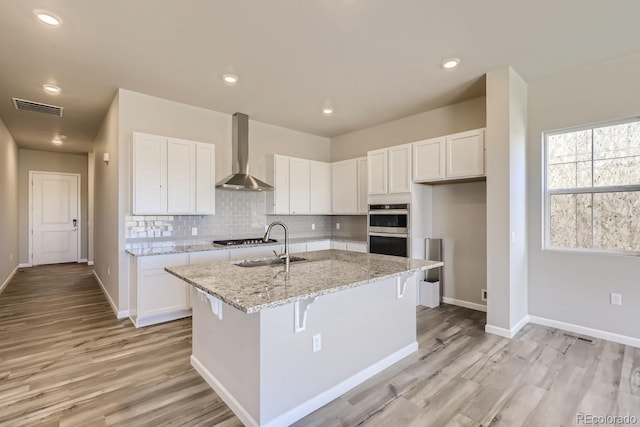 kitchen with a kitchen island with sink, white cabinets, light stone countertops, and wall chimney range hood