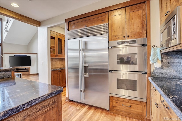 kitchen featuring vaulted ceiling with beams, light hardwood / wood-style flooring, backsplash, built in appliances, and dark stone counters