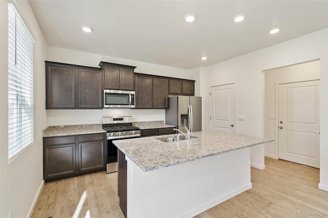 13467 Valentia St, Thornton, CO 80602 - kitchen with appliances with stainless steel finishes, dark brown cabinetry, a kitchen island with sink, light wood finished floors, and light stone countertops