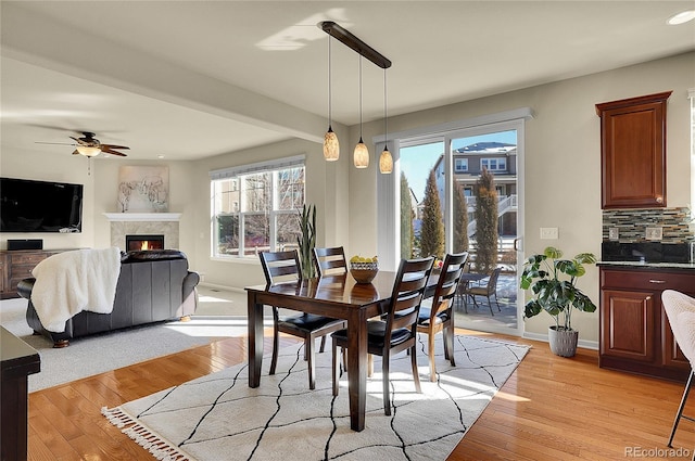 dining room featuring baseboards, light wood-style floors, a warm lit fireplace, and a ceiling fan