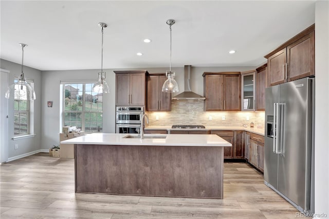 kitchen featuring wall chimney exhaust hood, a kitchen island with sink, light countertops, appliances with stainless steel finishes, and decorative backsplash