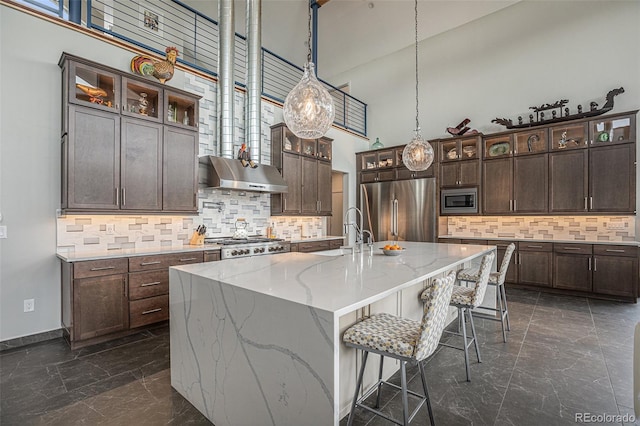 kitchen with appliances with stainless steel finishes, ventilation hood, a center island with sink, and a high ceiling